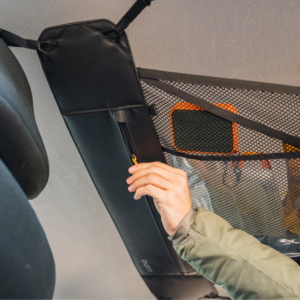 A person in a green jacket is using a zipper to close a black mesh storage net attached to a ceiling. The net appears to be part of a vehicle's interior storage solution. The image focuses on the person's hand and the storage net.