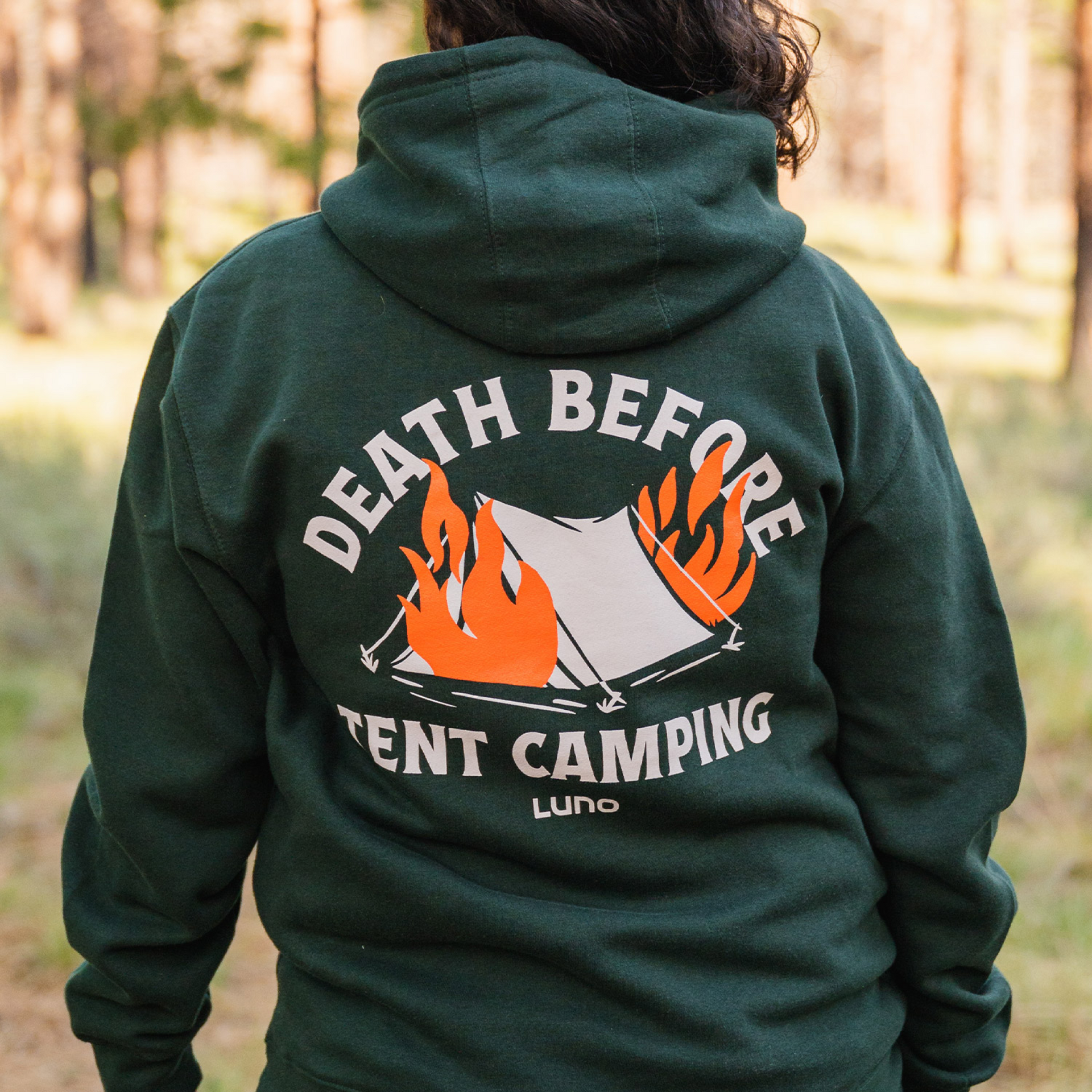 A person wearing the Luno HQ Death Before Tent Camping Hoodie, which showcases a tent on fire and the text "DEATH BEFORE TENT CAMPING," stays cozy in the outdoor, forested area thanks to its ultra-soft cotton and polyester blend.