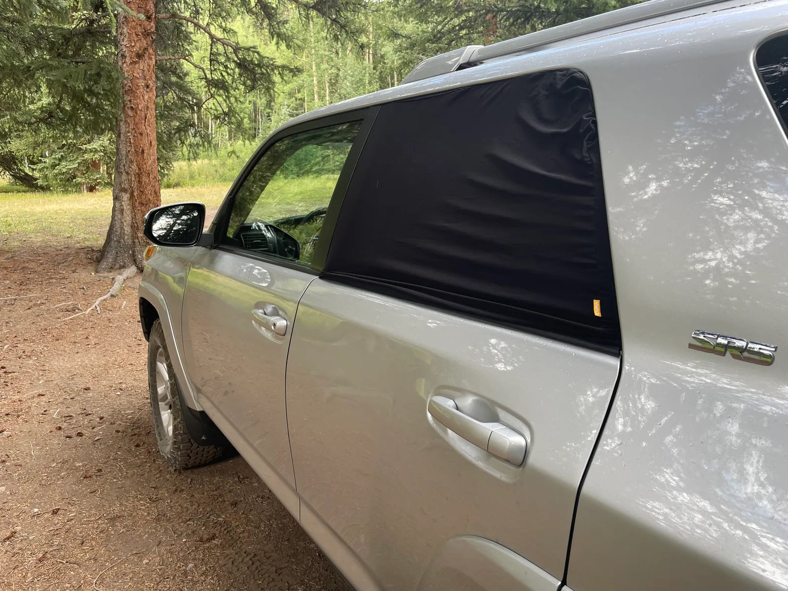 A silver SUV is parked in a wooded area. The rear window on the driver's side is covered with a black window shade. The side mirror and part of the vehicle's roof are visible. Tall green trees and grass can be seen in the background.