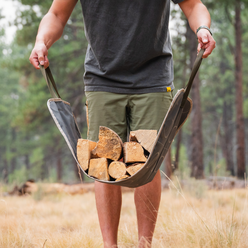 A person wearing a gray t-shirt and green shorts is holding an eco-friendly accessory—a Luno HQ Firewood Carrier Utility Mat filled with chopped firewood. The durable fabric carrier is stretched between their hands, and they are standing in a grassy outdoor area with trees in the background.