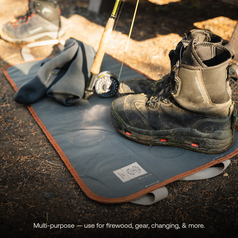 A pair of sturdy black hiking boots rest on a lightweight, grey Luno HQ Firewood Carrier Utility Mat made from durable fabric. Beside the boots are a rolled-up garment and a fishing rod. The text at the bottom of the image reads, "Multi-purpose – use for firewood, gear, changing, & more. This eco-friendly accessory is perfect for all your needs.