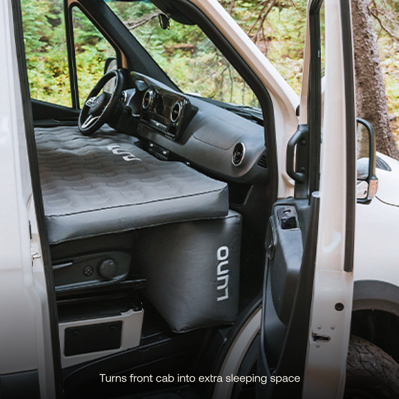 A van with the front door open reveals a Luno® AIR Van Front Cab Mattress made of durable fabric, placed over the front seats to convert the area into an extra sleeping space. The dashboard and steering wheel are visible. Text at the bottom reads, "Turns front cab into extra sleeping space.