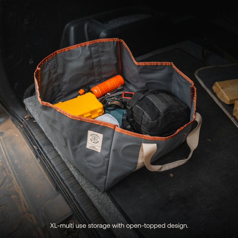 A Gear Tote | 50L from Luno HQ, featuring a large, open-topped design, is placed in the back of a vehicle. This versatile storage solution holds various items including a yellow case, an orange flashlight, a black bag, and other tools. Text at the bottom reads: "XL-multi use storage with open-topped design.
