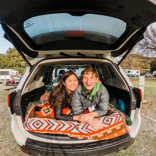 A smiling woman and man lie in the back of an open hatchback car, which is parked outdoors. They are resting on a brightly patterned blanket with the car's trunk open, surrounded by a grassy area with scattered trees and vehicles in the background.