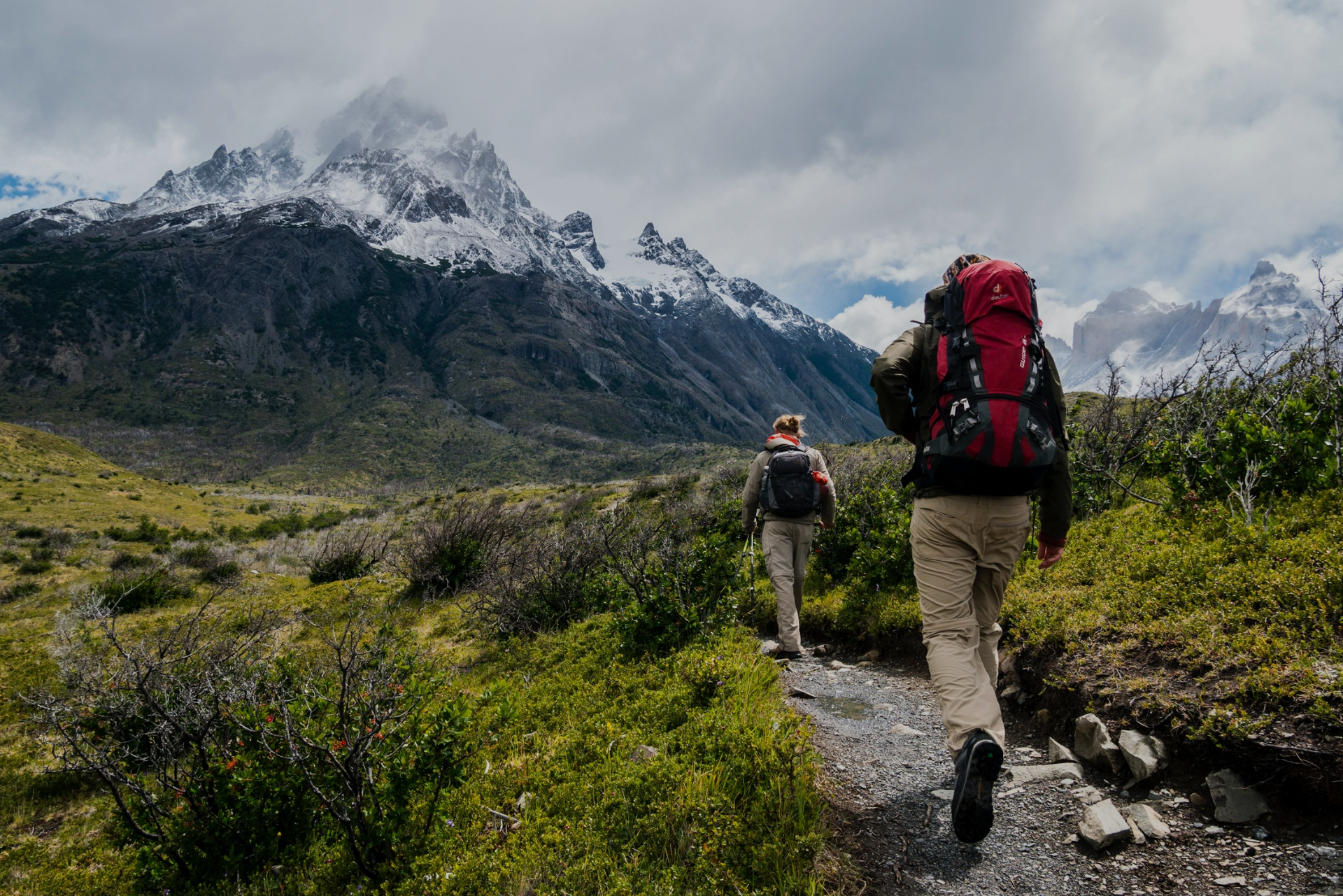 Two hikers with backpacks walk along a trail through a green valley towards towering, snow-capped mountains under a cloudy sky. The landscape features rugged terrain and sparse vegetation. The hikers appear focused on their journey.