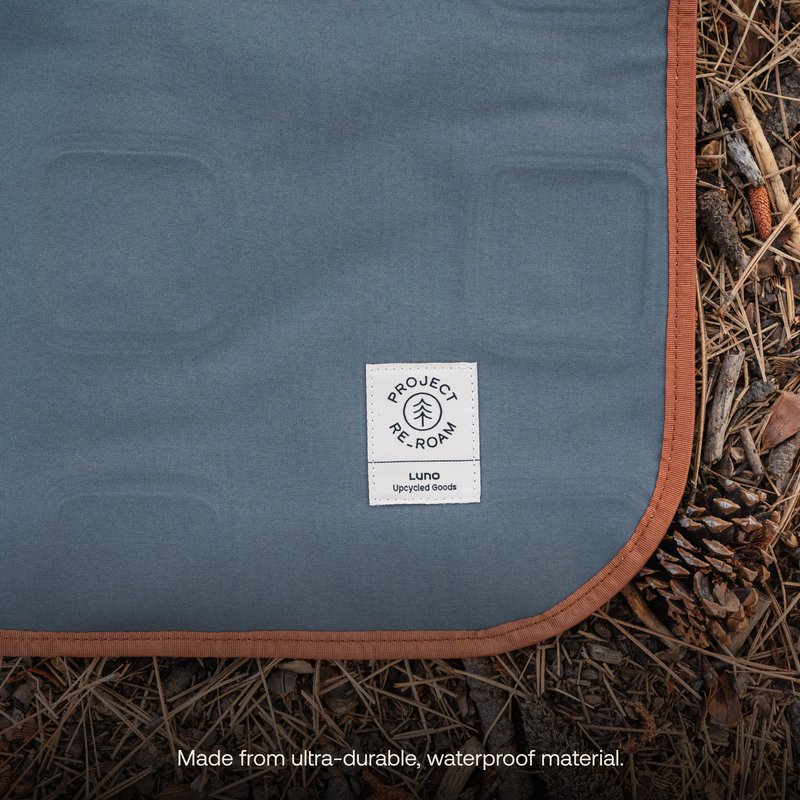 A close-up of a blue outdoor blanket with a brown edge lies on forest ground covered with dried leaves and pine needles. A white label on the blanket reads "Luno HQ Upcycled Goods." The caption below states, "Made from ultra-durable, waterproof material, ideal as an eco-friendly Large Outdoor Ground Mat.