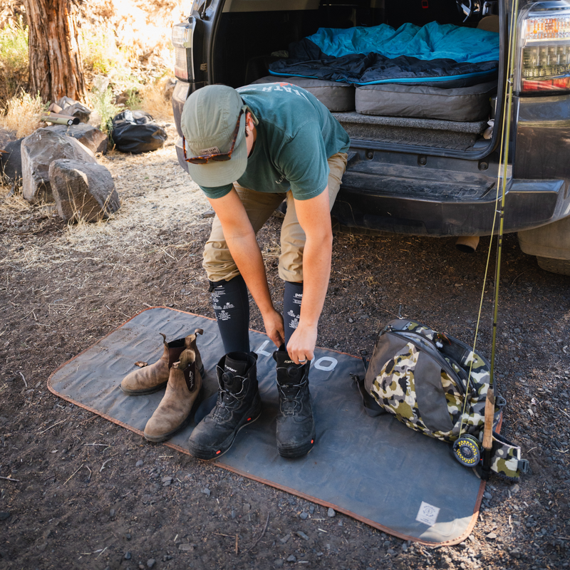 A person in outdoor clothing is putting on hiking boots next to a car with an open trunk. Inside, the trunk showcases a sleeping setup featuring upcycled mattresses. The person stands on the Luno HQ Large Outdoor Ground Mat, with additional gear such as a backpack and fishing rod nearby on the ground.
