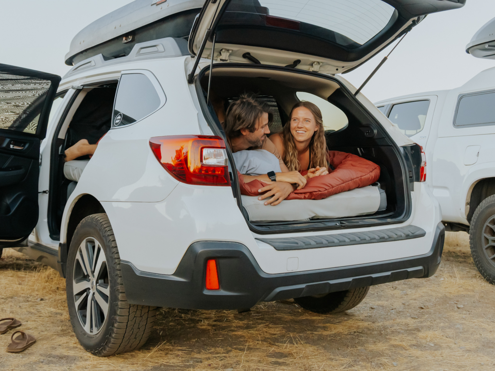 Two people lie in the back of a white SUV with the trunk open. They look at each other and smile, surrounded by pillows and blankets, indicating they are camping. The SUV is parked on dry, grassy ground with another vehicle in the background.