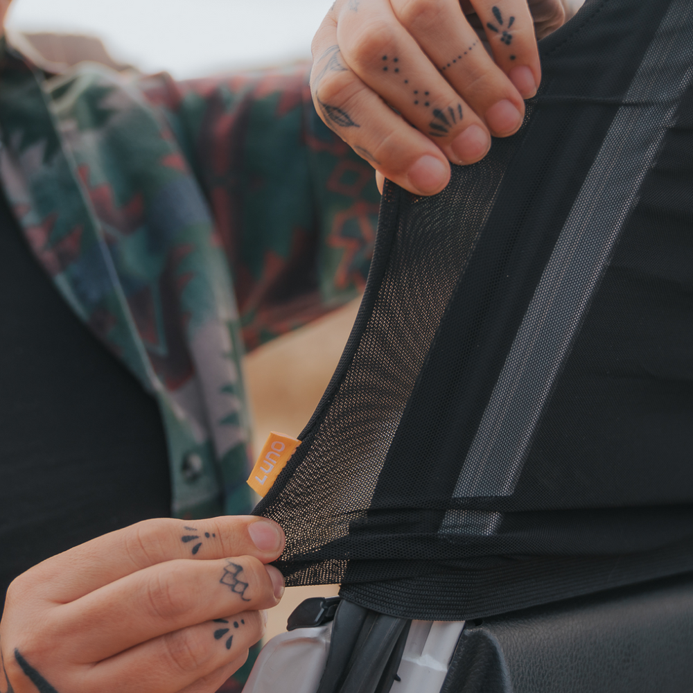 A person with tattoos on their fingers stretches a black mesh material attached to a car window. They are holding an orange tag labeled "Luno." The person is wearing a colorful patterned fabric and a black top. The background is indistinct and blurred.