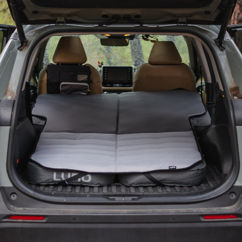 A vehicle's rear cargo area with the back seats folded down and a Luno® Memory Foam Topper laid out, creating a flat sleeping surface. The car campers are parked in a wooded area, with trees visible in the background through the open trunk.