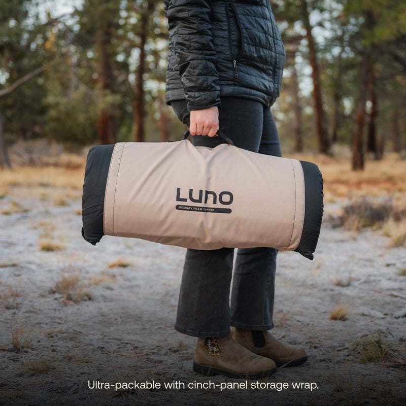 Wearing a black jacket and brown boots, a person carries a rolled-up Luno HQ Memory Foam Topper with a cinch-panel storage wrap, ideal for car camping. The grassy outdoor setting with trees enhances the adventure-ready vibe.