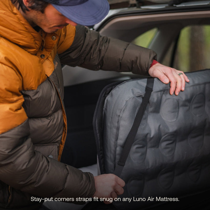 A person wearing a brown and green jacket adjusts straps on a pre-owned Memory Foam Topper by Luno HQ in the back of a vehicle. This comfy mattress features stay-put corner straps, making it perfect for car camping adventures.