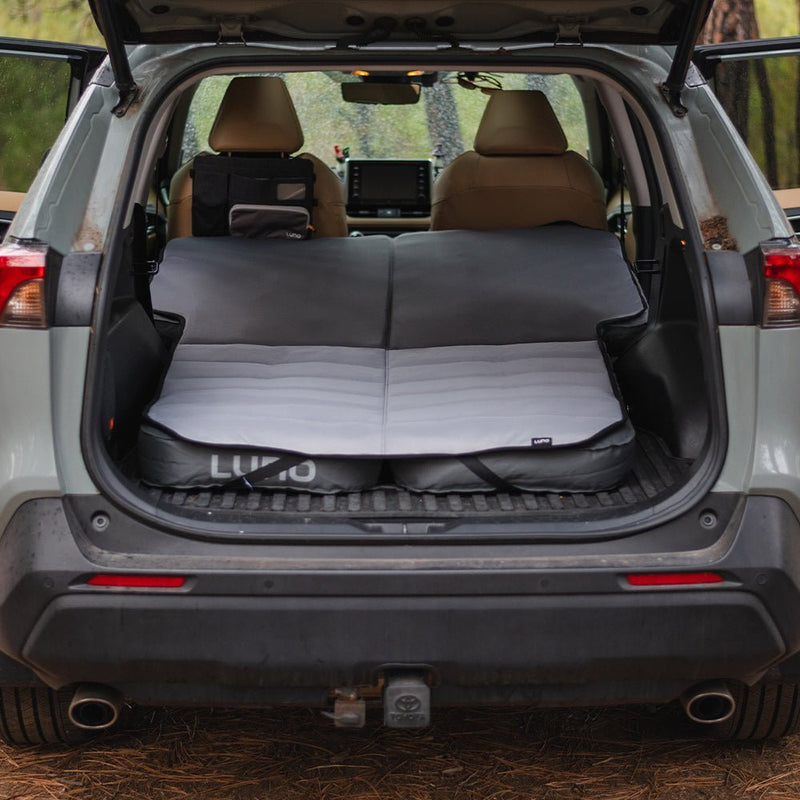 An SUV with its rear hatch open showcases a neatly arranged Luno HQ Memory Foam Topper (Pre-Owned) for optimal comfort. Surrounded by trees, it presents an ideal car camping scene. The front seats are in view, along with a navigation screen on the dashboard.
