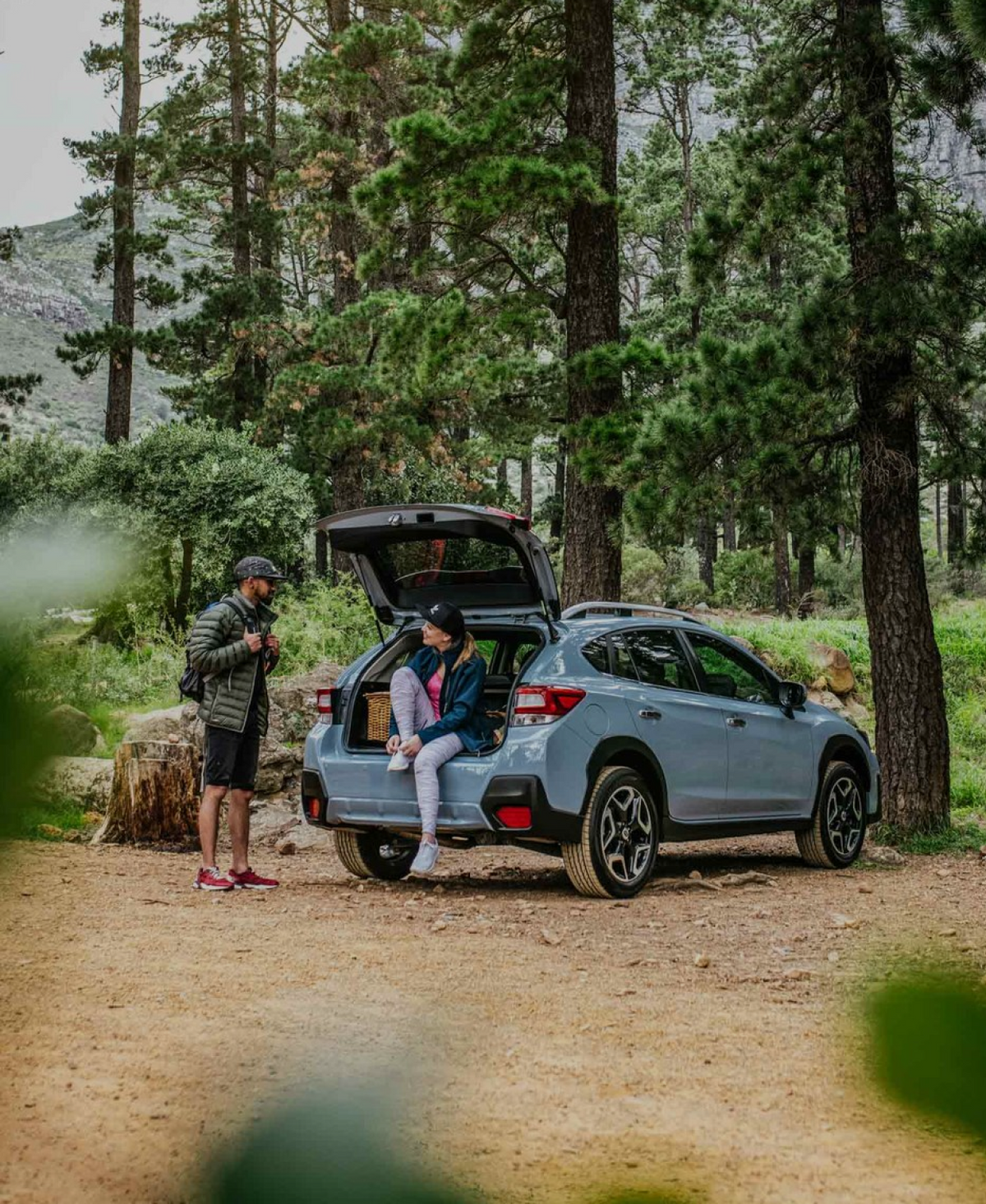 Two people in an outdoor setting are standing by a light blue SUV with its trunk open. One person is sitting on the trunk edge, the other stands nearby holding trekking poles. The scene is surrounded by tall trees and greenery, suggesting a forested area.