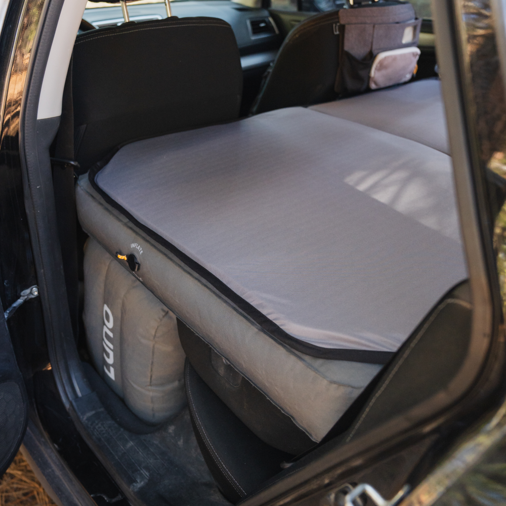 A car interior with the rear seats folded down and a grey inflatable mattress laid out for car camping. A storage pouch is attached to the back of the front passenger seat. The setup is designed for a comfortable sleeping space inside the vehicle.