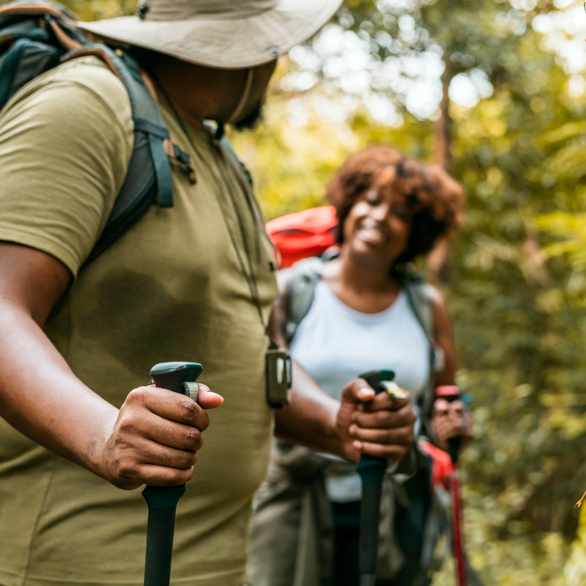 Two people are hiking in a forest, wearing casual outdoor attire and carrying backpacks. The person in the foreground wears a wide-brimmed hat and holds walking poles, while the person in the background is smiling and also holding walking poles. .