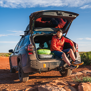 A person in an orange jacket and cap sits at the open rear of a parked SUV on a dirt road. The vehicle is loaded with camping gear, and the sky is clear and blue. The surrounding landscape is dry and rocky, suggesting a desert or arid area.