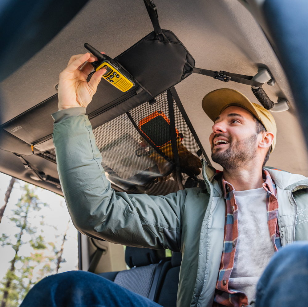 A man inside a vehicle smiles as he reaches up to secure a yellow handheld radio into an overhead mesh storage net. He is wearing a green jacket, plaid shirt, and beige cap. The background shows a glimpse of trees outside the vehicle.
