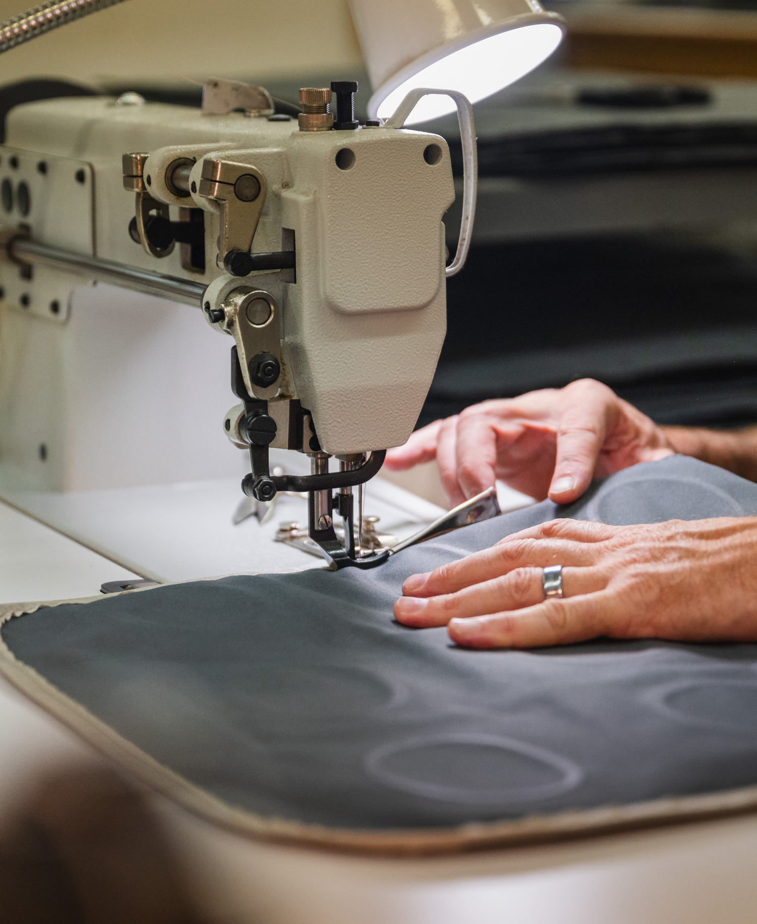 A person with a ring on their finger uses an industrial sewing machine to stitch a piece of dark fabric. The workspace is well-lit with a lamp attached to the sewing machine.