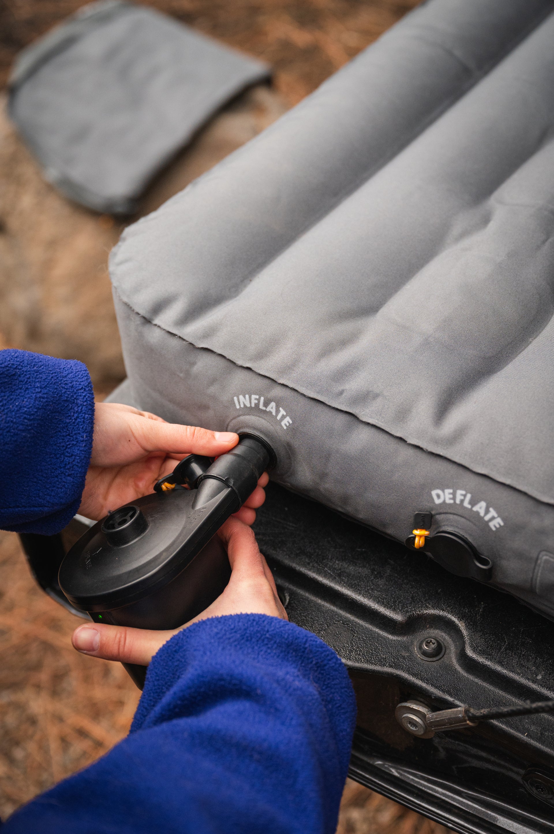 A person in a blue fleece jacket uses an electric air pump to inflate a gray air mattress. The mattress has visible labels for "INFLATE" and "DEFLATE," and it is positioned on the edge of a truck tailgate in an outdoor setting.