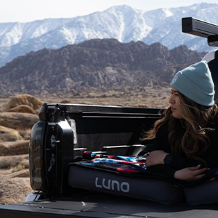 A person in a light blue beanie reclines in the back of a truck with a Luno mattress, set against a backdrop of rugged mountains and rocky terrain. The scene suggests a camping or outdoor adventure.