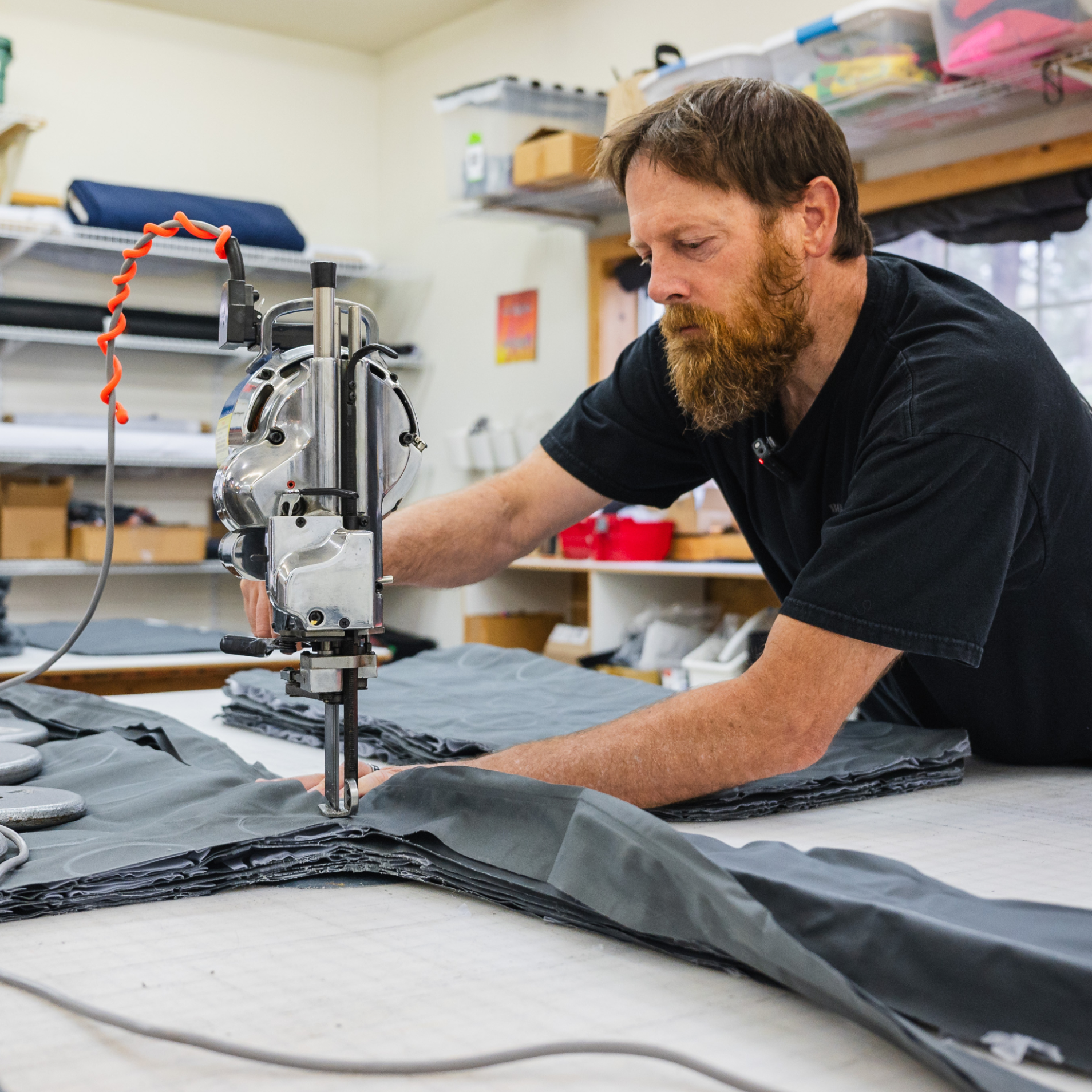 A bearded man in a black T-shirt operates a fabric cutting machine on a large piece of gray fabric in a well-organized workshop. Shelves with various supplies are in the background. The man is focused on his task.