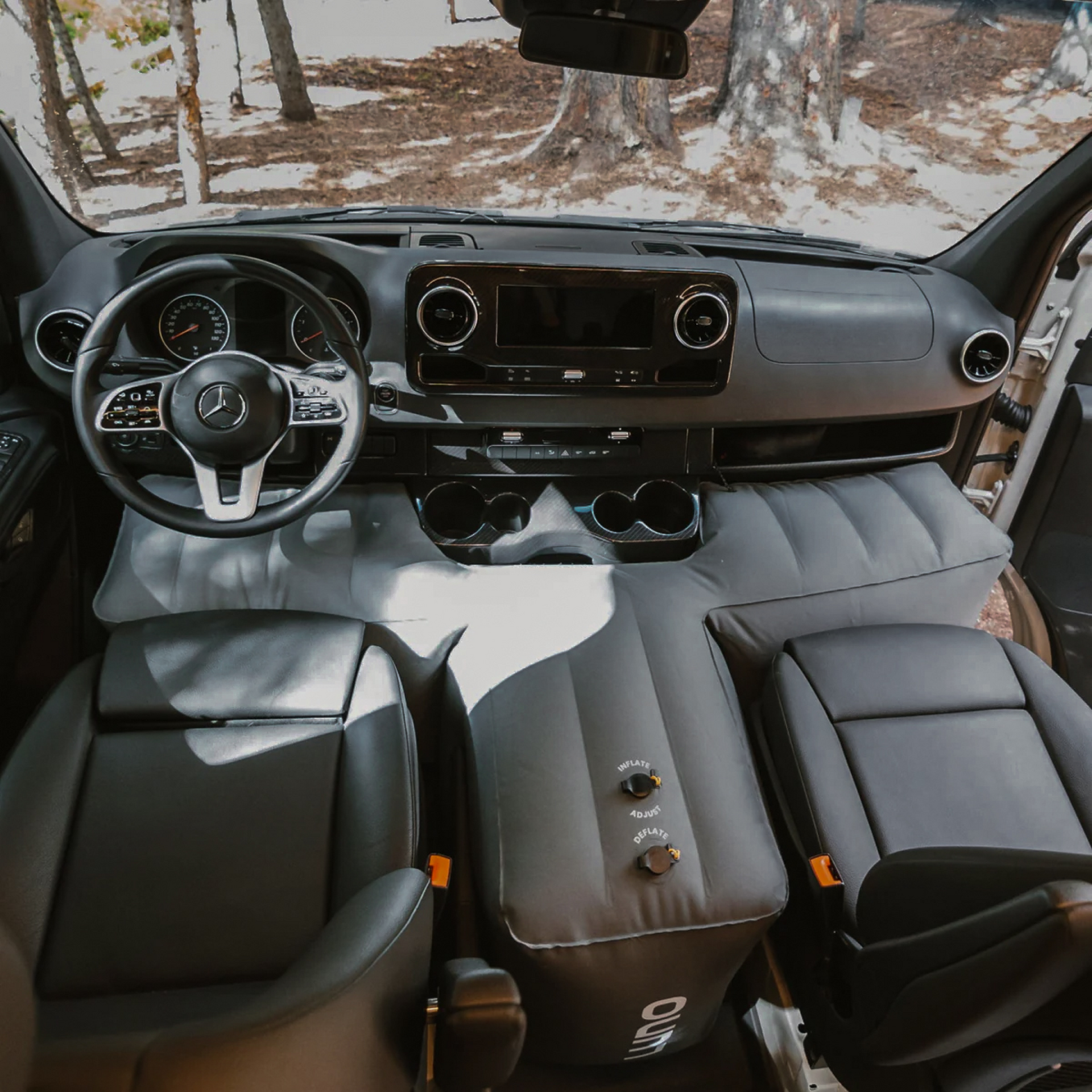 Interior view of a modern camper van, featuring a sleek black dashboard with a Mercedes-Benz logo on the steering wheel. The front seats are facing inward, with a grey inflatable **Luno HQ Van Front Cab T-Shaped Space Filler (Pre-Owned)** extending between them, creating additional space for lounging or sleeping.