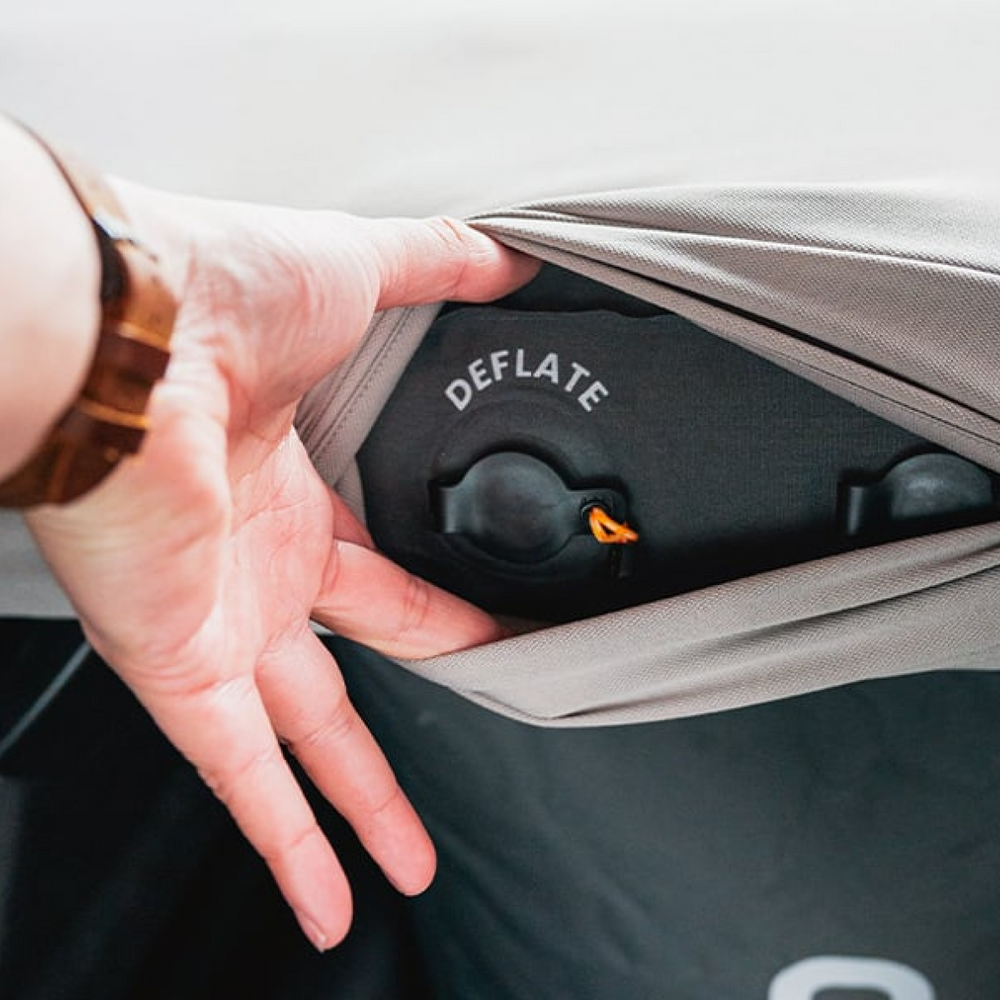 A person is pulling back a piece of fabric to reveal a black valve with the word "DEFLATE" written near it. The valve appears to be part of an inflatable object, and the person’s wrist has a brown watch.