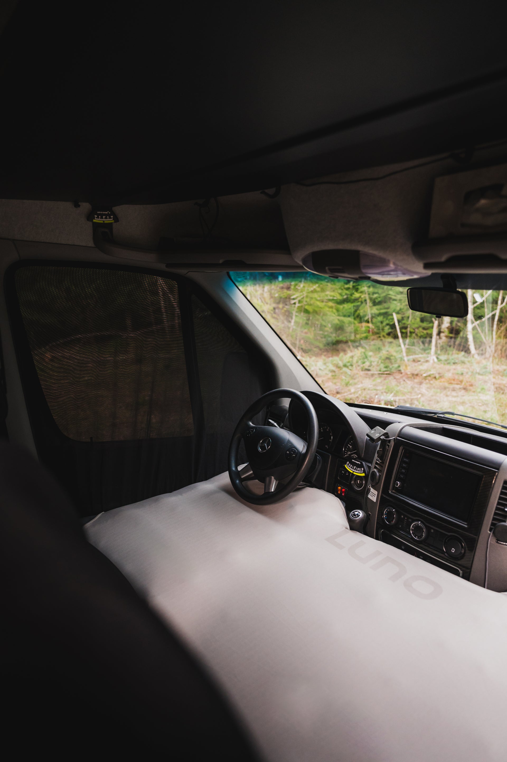 Interior of a camper van showing a driver's seat, steering wheel, and dashboard with a black and gray color scheme. The van has a large Luno-branded inflatable mattress covering the front seats, with a leafy forest visible through the windows.