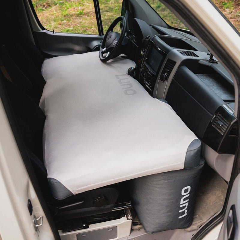 The interior of a van features a pre-owned air van front cab mattress fitted sheet, extending from the dashboard to the front seats. "Luno HQ" is visible on both the sheet and a bag beneath it. The well-lit scene, with greenery visible through the windshield, highlights USA shipping.