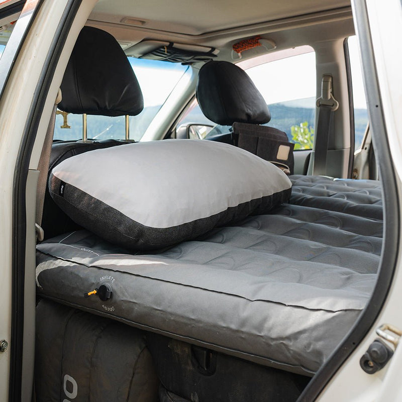 Inside the car's interior, a gray inflatable mattress spans the back seats, adorned with a matching luxury pillow. The front seats are visible as sunlight streams through the windows. Experience on-the-go comfort with our Luno HQ Packable Camp Pillow (Pre-Owned), now easily accessible with convenient shipping across the USA.
