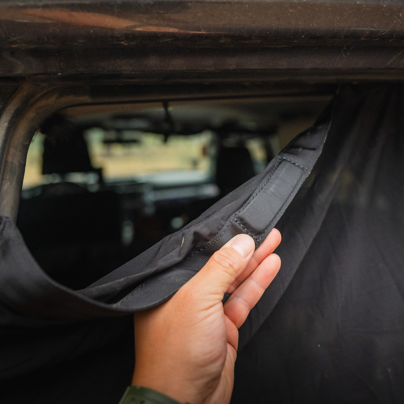 A person's hand is holding and adjusting a Luno® 4Runner Trunk Window Screen, which features a black fabric curtain secured with a Velcro strip inside a vehicle. This essential car camping accessory covers the window, with the interior of the vehicle slightly visible in the background.