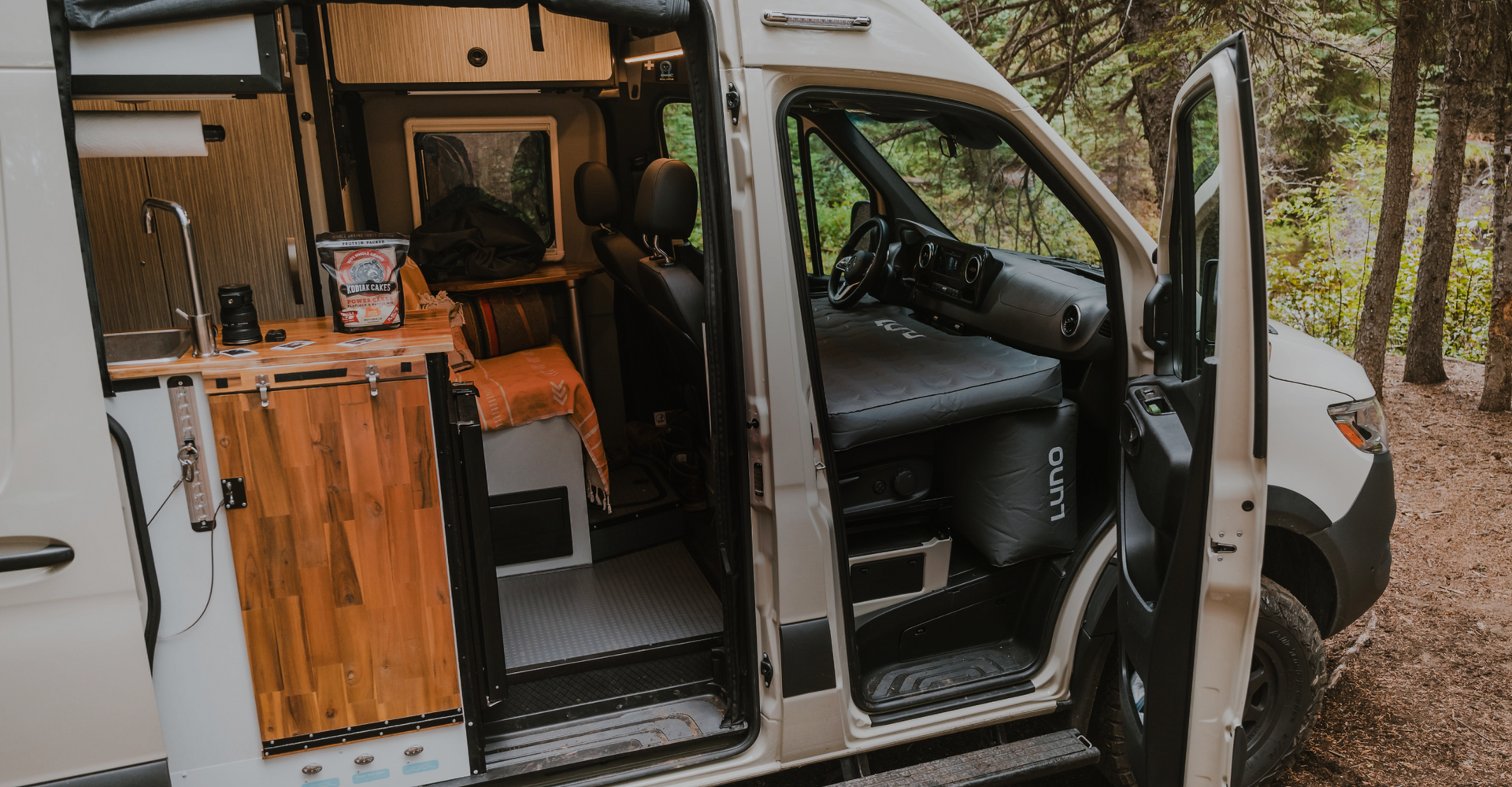 A white camper van with open sliding doors reveals a compact, neatly organized interior. The van is equipped with wooden cabinetry, a small countertop, and a driver's area. The surrounding environment includes trees and greenery, indicating a forest setting.