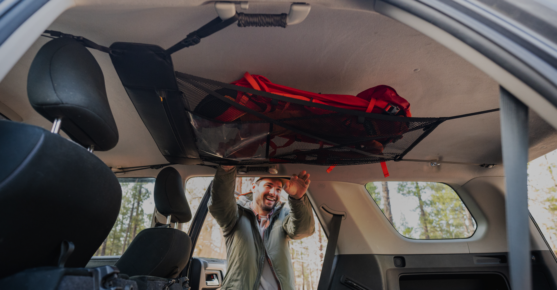 A person arranges a red backpack in an overhead storage net inside a vehicle. The interior of the vehicle has black and grey seats, and the rear door is open. Trees are visible through the windows, indicating an outdoor location.