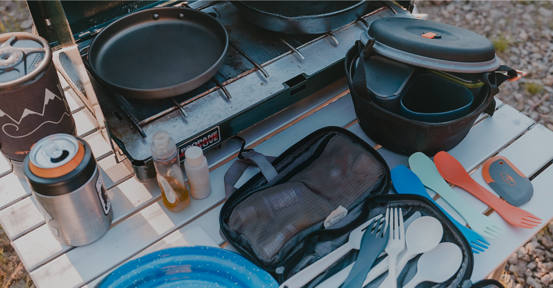 A camping setup on a white table showing a portable gas stove with pans, a travel mug, small condiment bottles, utensils, a cook set with bowls, plates, and cutlery, along with a zippered pouch containing cooking and cleaning items. The ground is gravelly.