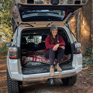 A person wearing a beanie and maroon sweatshirt sits on the edge of an SUV's open trunk, which is set up for camping with a cozy setup including a blanket. The vehicle is parked in a wooded area with tall trees in the background.