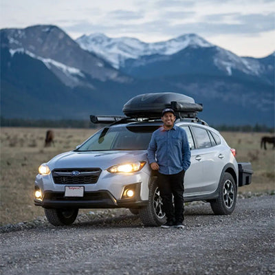 A man stands beside a silver Subaru SUV on a gravel road with mountainous terrain in the background during dusk. The SUV has a roof cargo box and the headlights are on. Several horses can be seen grazing in the field behind him.