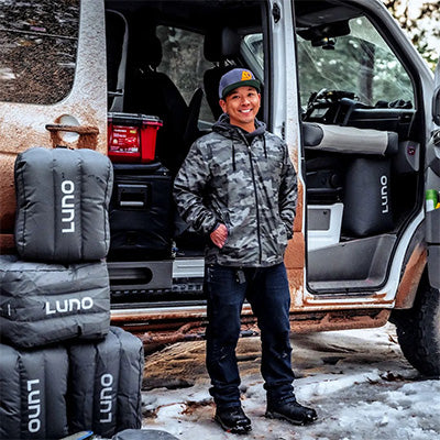 A person dressed in a camo jacket, black pants, and a beanie stands beside a van in a snowy outdoor setting. The van is partially covered in mud and has camping gear labeled "LUNO" stacked next to it.