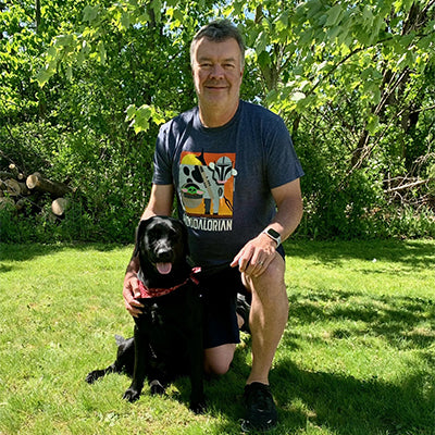 A man kneels on the grass next to a black dog in a leafy, green park. The man is smiling and wearing a "Mandalorian" T-shirt with shorts. The dog has a red bandana around its neck and is sitting calmly beside the man. Sunlight filters through the trees behind them.