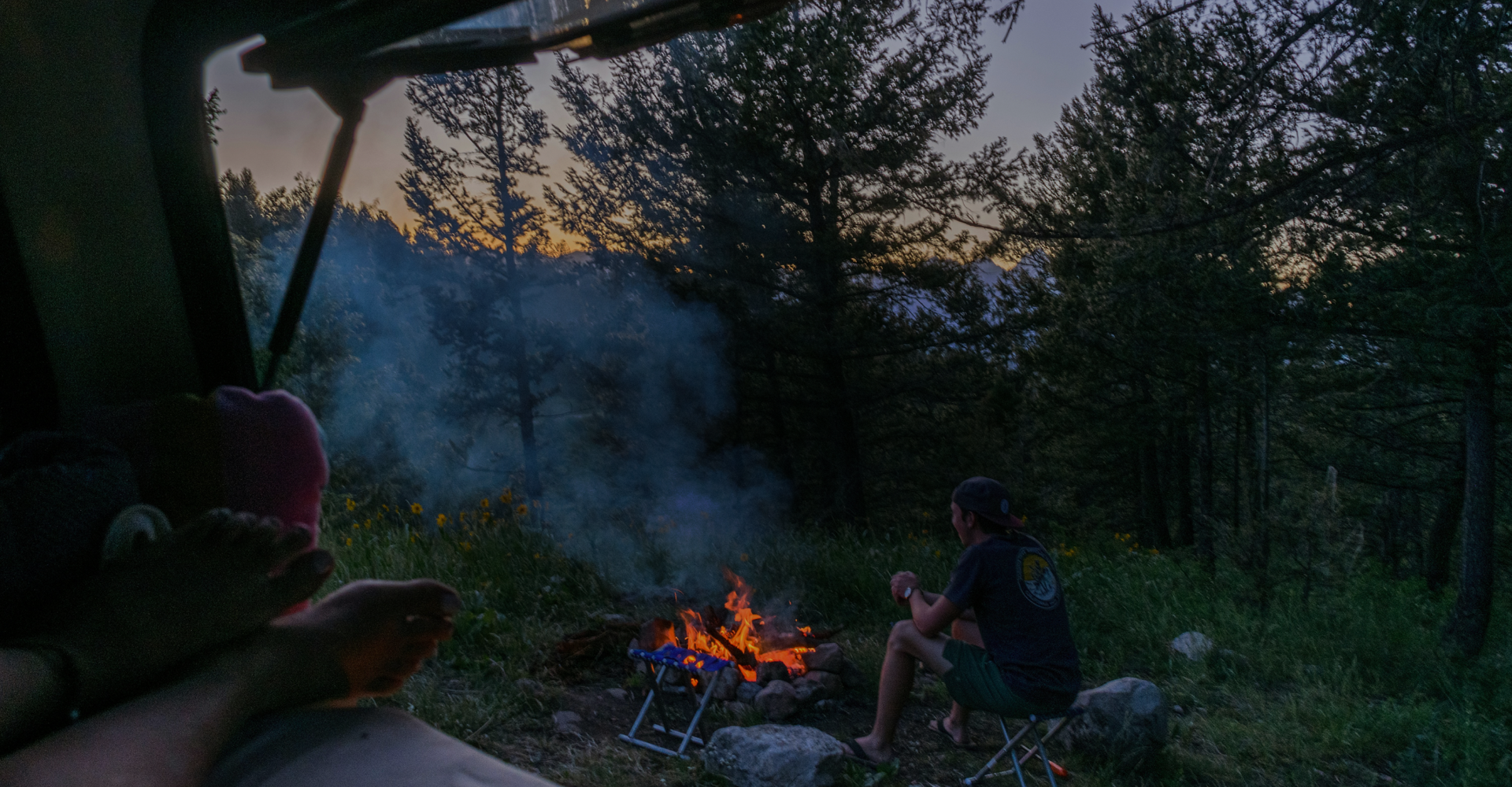 Person sitting on a folding chair by a campfire in a forested area during twilight, viewed from inside a vehicle. Smoke rises from the fire, and the scene is surrounded by tall trees. One person's feet are visible in the foreground, resting inside the vehicle.
