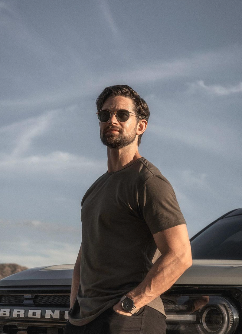 A man with dark hair and a beard stands outdoors in front of a vehicle, wearing sunglasses, a dark green t-shirt, and a watch. He has his hands in his pockets and looks off into the distance under a clear sky.