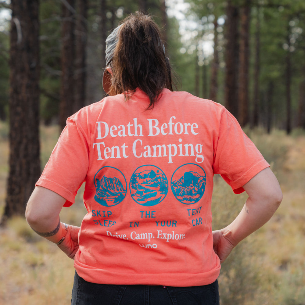 Outdoors in a forest, someone wears the Death Before Tent Camping Unisex T-Shirt featuring bold white text on the back: “Death Before Tent Camping. Skip the tent, sleep in your car. Drive. Camp. Explore.”—ideal for fans of the Luno lifestyle.