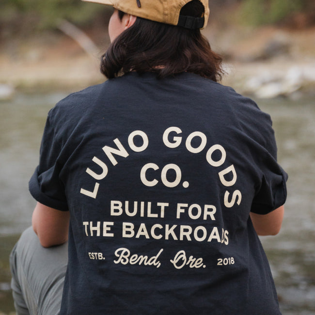 A person wearing a classic fit beige cap and a dark blue Luno Goods Camping T-Shirt by Luno HQ sits by a riverside. The ultra-soft cotton back of the t-shirt displays: "Luno Goods Co. Built for the Backroads. Estb. Bend, Ore. 2018." The natural background features blurred greenery and water.