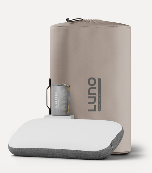 A light beige Luno camping pillow, featuring a compact, rectangular design with a white and gray color scheme, is displayed in front of its matching storage bag. The storage bag includes a smaller, cylindrical item also marked with the Luno logo.