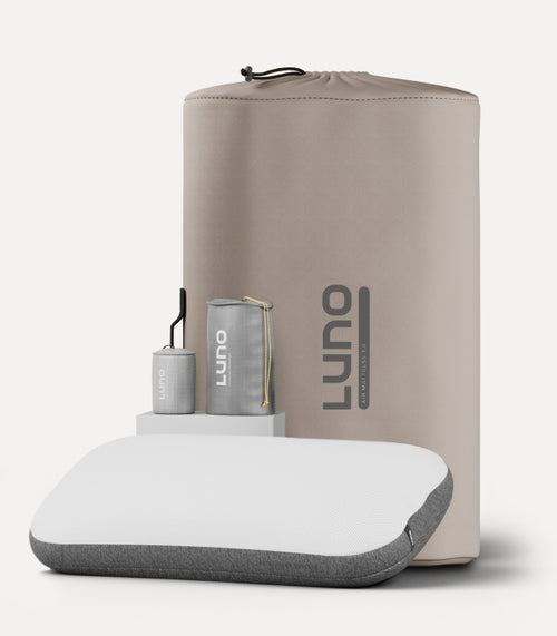 A memory foam pillow with a grey base and white top is displayed in front of a beige carrying case labeled "Luno." Two small, cylindrical travel-sized pillowcases and a rectangular case are also shown, highlighting the pillow's portable and compact design.