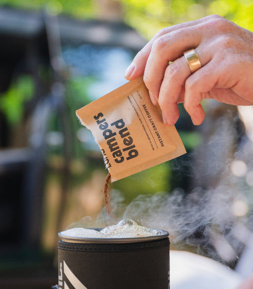 A hand with a gold ring pours a powdered coffee mix labeled "campers blend" from a torn packet into a steaming cup, with an outdoor, blurred background hinting at a camping scene.