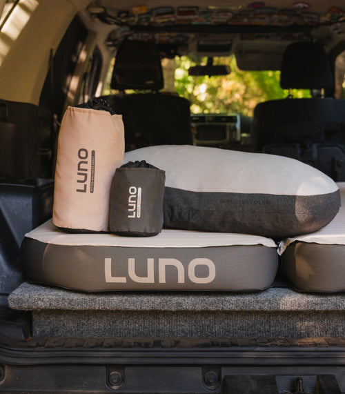 A neatly organized car trunk features a gray and black inflatable mattress with the brand name "Luno" displayed prominently. Two rolled-up Luno sleeping bags, one beige and one charcoal gray, are placed on top of the partially inflated mattress against a backdrop of lush greenery.