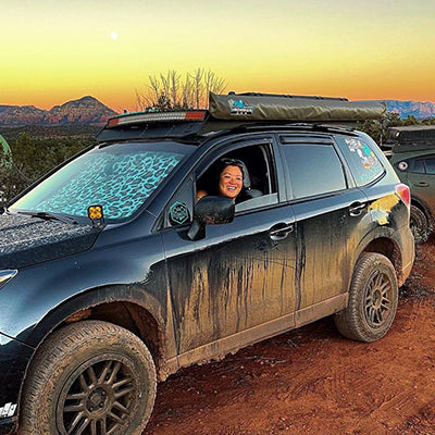 A person smiling and sticking their head out of the window of a muddy SUV with off-road equipment. The vehicle is on a dirt road with mountains and a sunset in the background.