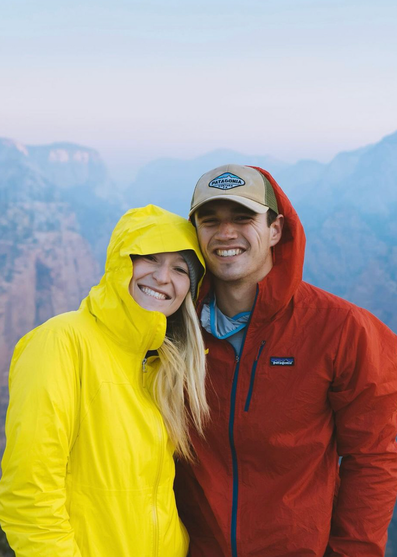 A smiling couple stands close together outdoors, with the woman on the left wearing a yellow jacket and the man on the right wearing a red jacket and a beige cap. They are against a backdrop of mountains shrouded in mist.