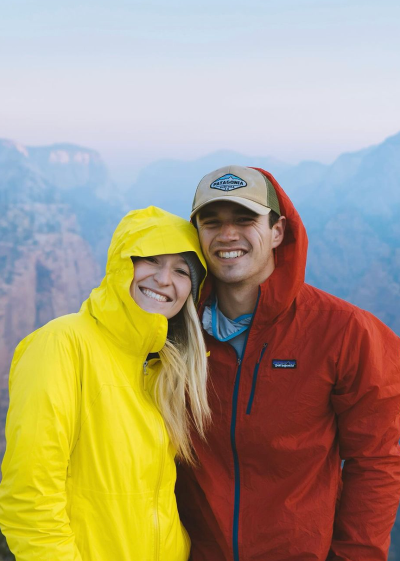 A smiling man and woman stand close together, wearing bright outdoor jackets; the man in red and the woman in yellow. The man wears a light cap and both have hoods down, against a background of misty mountains.